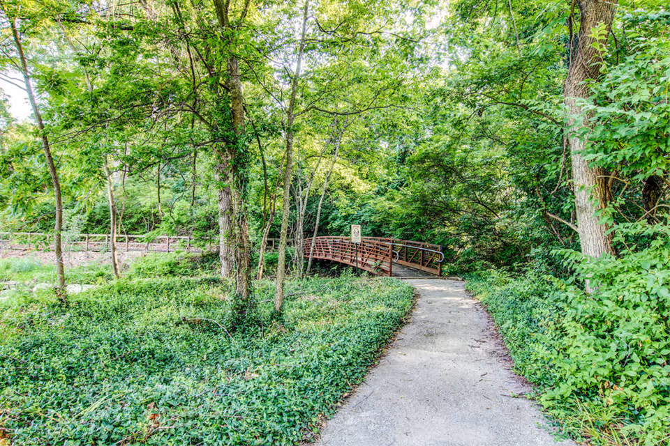 A paved walking path surrounded by dense green foliage and trees leading to a small wooden bridge in a lush outdoor setting.