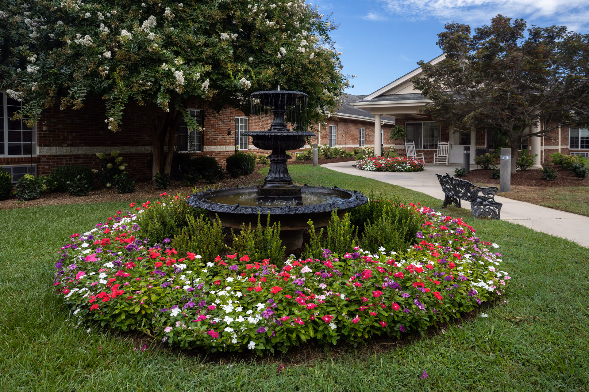A landscaped garden area at Oak Hill Assisted Living Community featuring a black tiered water fountain surrounded by colorful flowers and green shrubs. There is a paved walkway with a black metal bench and a brick building with white-trimmed windows and a covered porch with white rocking chairs in the background. Trees with green and reddish leaves frame the scene under a partly cloudy blue sky.