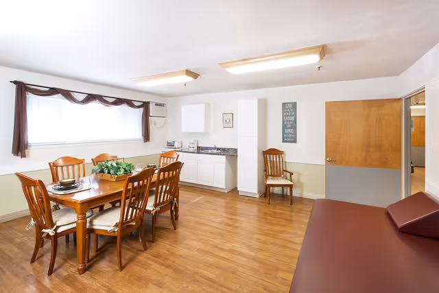 Well-lit common dining area with a wooden table and chairs, a small kitchenette, and additional seating.