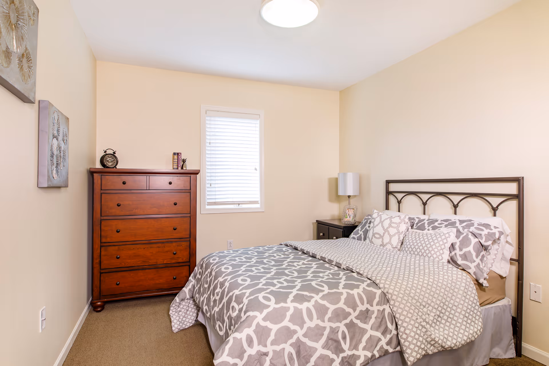 A small bedroom with a patterned bed and metal headboard, wooden chest of drawers, nightstand with a lamp, and a window.