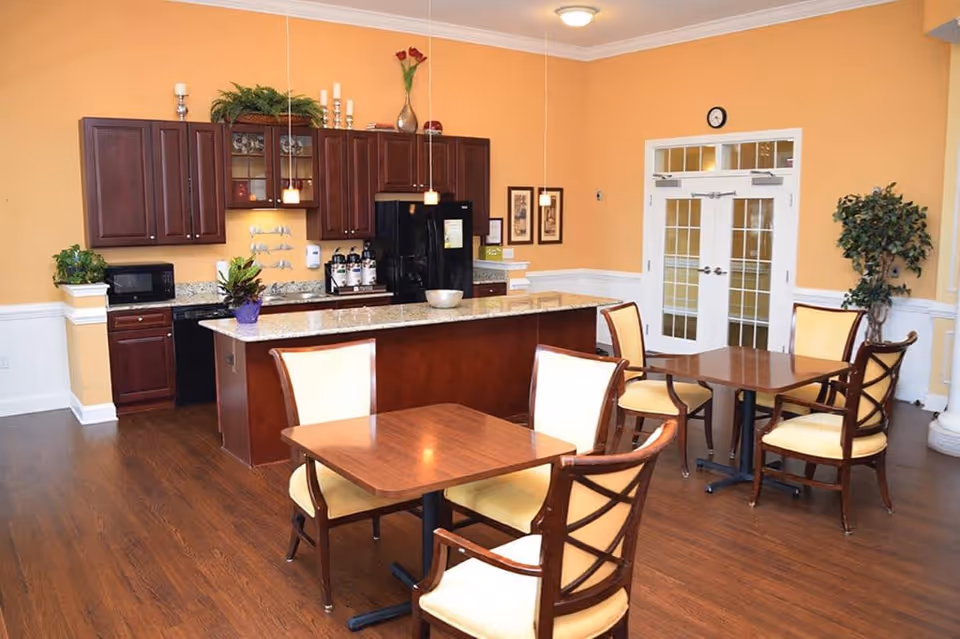 A bright dining area with wooden floors and yellow walls featuring a kitchen with dark wood cabinets, a granite countertop island, and appliances including a microwave and refrigerator. There are two wooden tables each surrounded by four cushioned chairs with wooden frames. The room has pendant lights hanging over the island, framed pictures on the wall, a clock above double glass doors, and potted plants for decoration.
