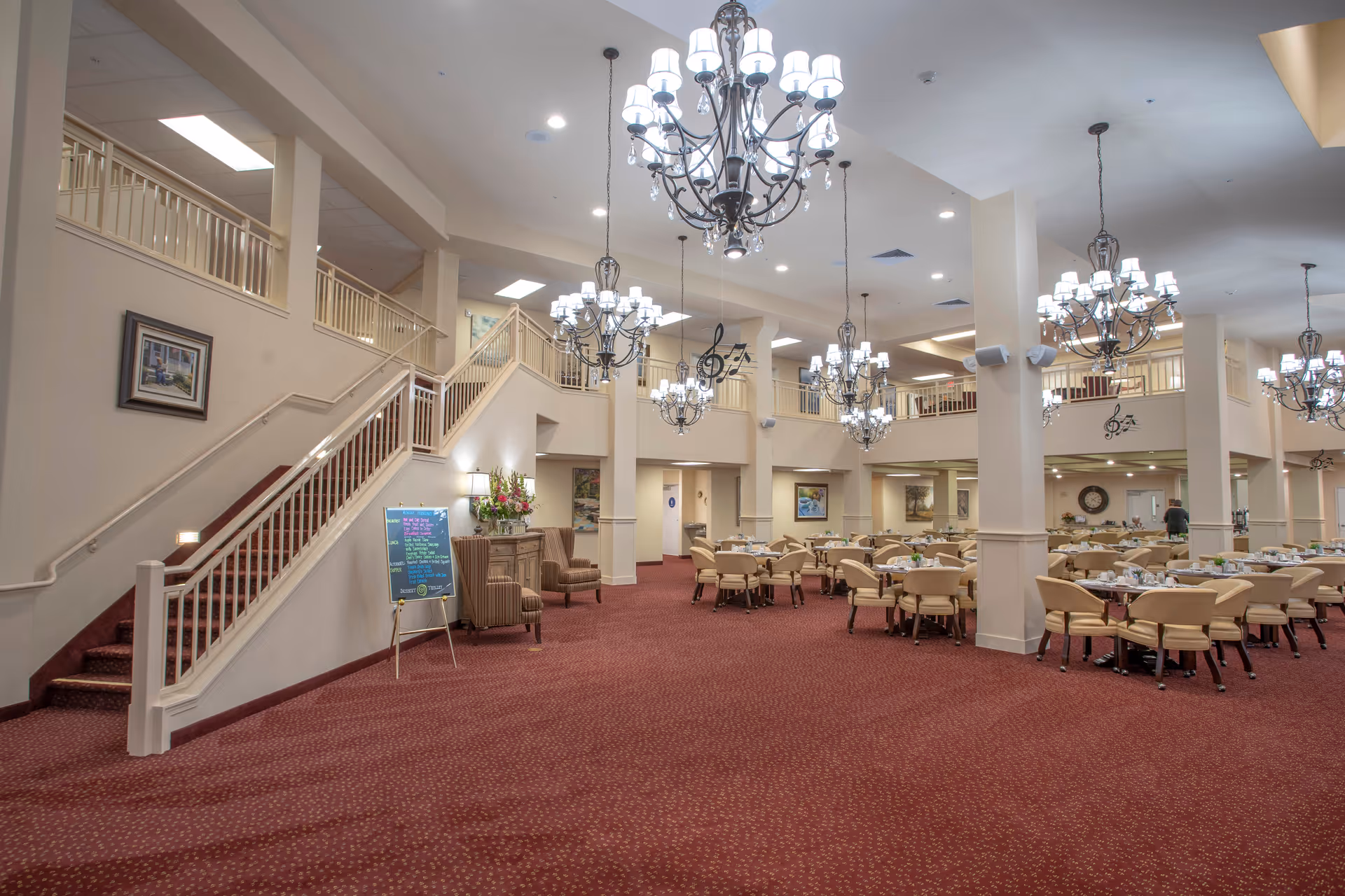 Spacious dining area in a retirement living facility with multiple round tables and chairs arranged neatly. The room features red carpet flooring, cream-colored walls, and several elegant chandeliers hanging from the ceiling. There is a staircase with a white railing on the left side, and the upper level has a balcony overlooking the dining area. Some framed artwork and decorative elements are visible on the walls.