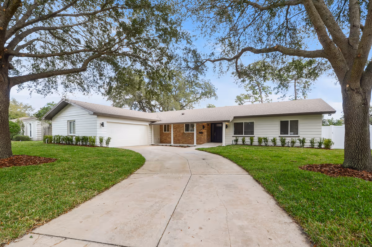 Single-story ranch-style home front with a curved concrete driveway, manicured lawn, and large oak trees.