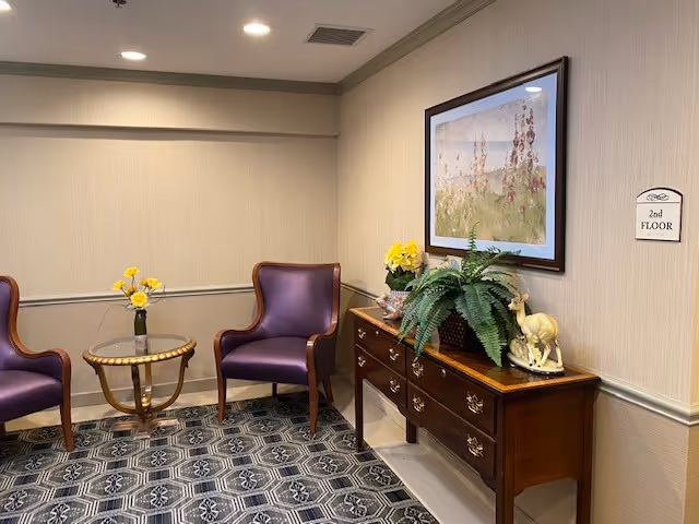 A small sitting area with two purple armchairs and a round glass-top table with a vase of yellow flowers. Next to the chairs is a wooden console table with drawers, decorated with a potted fern, yellow flowers, and a small deer figurine. A framed floral painting hangs on the wall above the console table. A sign on the wall indicates this is the 2nd floor.