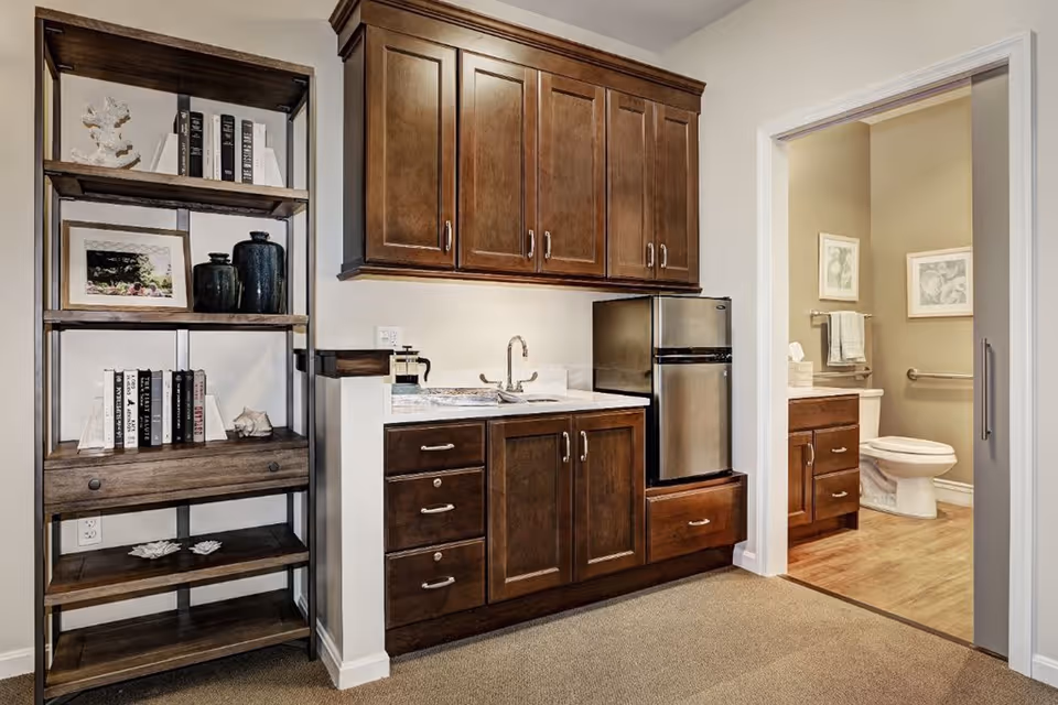Interior view of a senior living facility room featuring a wooden shelving unit with books and decorative items on the left, a kitchenette with dark wooden cabinets, a small sink, and a mini refrigerator in the center, and a bathroom with a toilet, vanity, and framed pictures visible through an open doorway on the right.