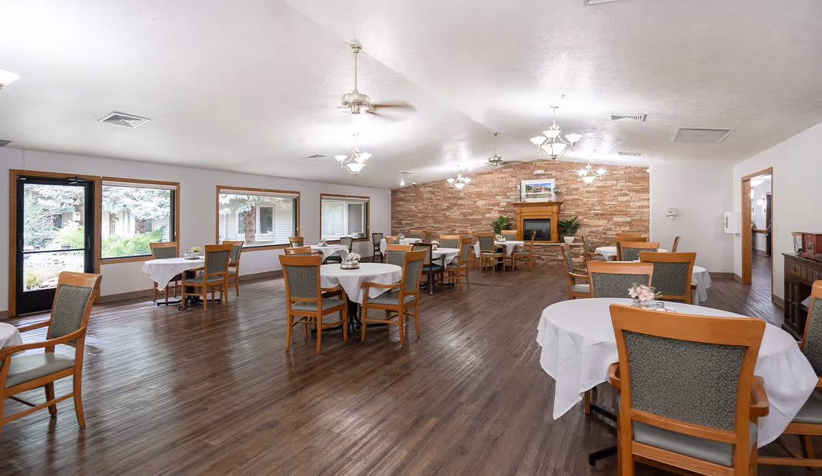 A spacious dining room with multiple round tables covered with white tablecloths and surrounded by wooden chairs with cushioned seats. The room has large windows letting in natural light, a wooden floor, ceiling fans, and a stone accent wall with a fireplace and framed artwork above it.