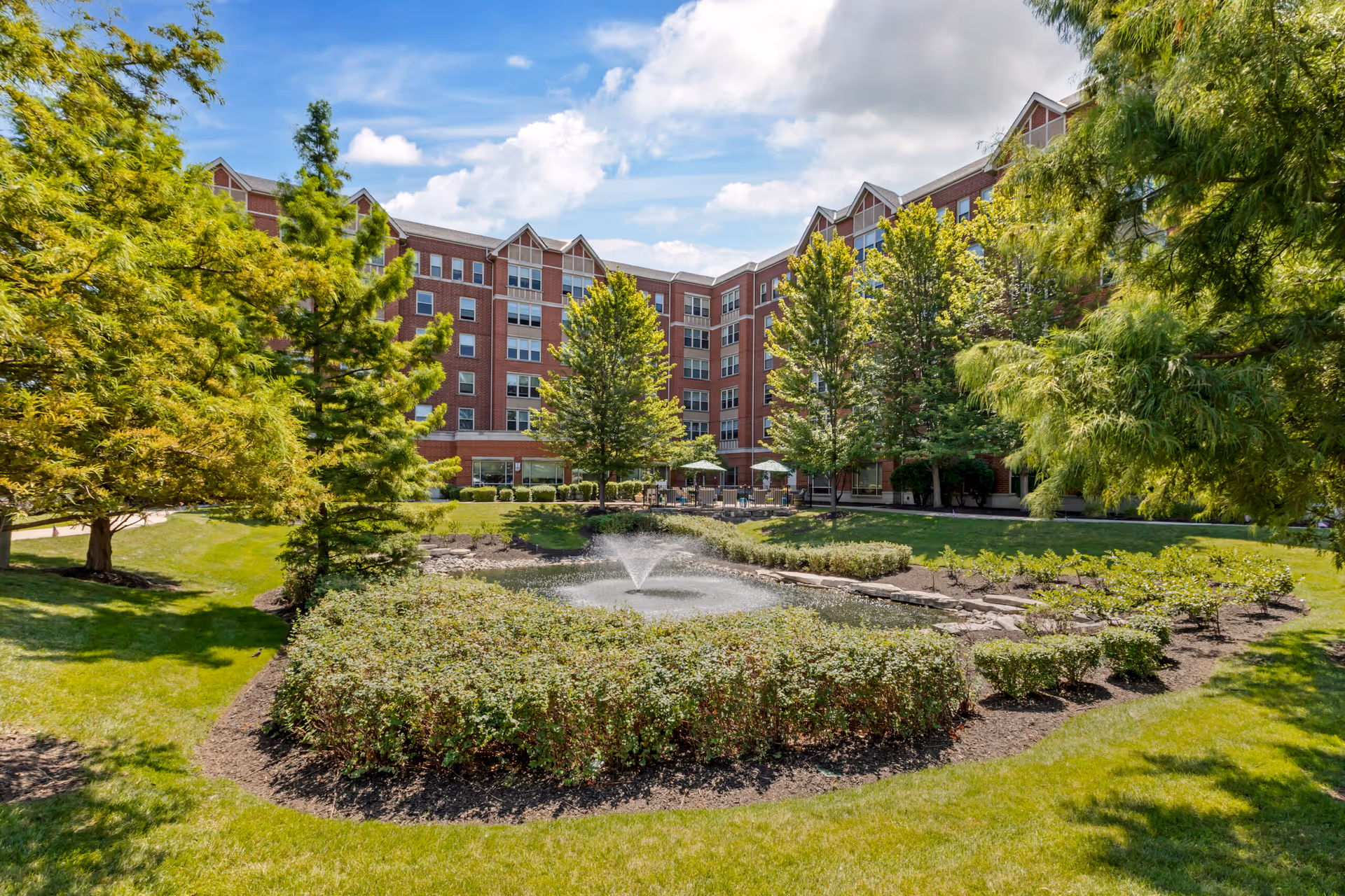 A landscaped outdoor area with a small pond featuring a water fountain in the center, surrounded by bushes and trees. In the background, there is a multi-story brick building with many windows under a partly cloudy blue sky.