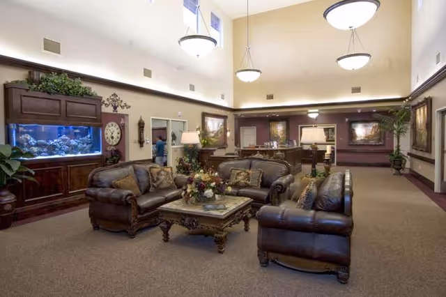 A spacious senior living facility common area with high ceilings and warm lighting. The room features three dark leather sofas arranged around an ornate coffee table with a floral centerpiece. To the left, there is a large built-in aquarium with colorful fish and a decorative clock on the wall. The walls are adorned with framed paintings and there are several potted plants adding greenery to the space. In the background, a reception desk is visible with a person standing nearby.