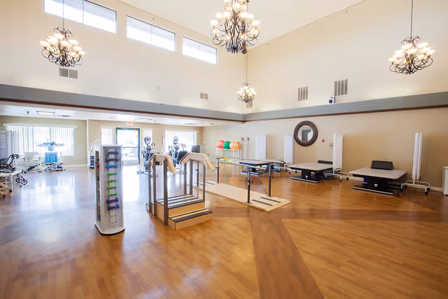 A spacious rehabilitation or physical therapy room with wooden floors, exercise equipment including parallel bars, steps, therapy tables, exercise balls, and weights. The room has high ceilings with multiple chandeliers and large windows letting in natural light.