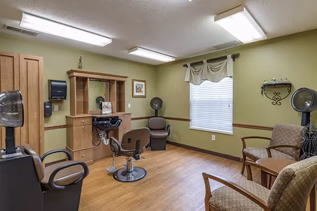 Interior of a senior living facility hair salon with salon chairs, hair dryers, a mirror, and a wooden cabinet against a light green wall. There are also two armchairs and a small table near a window with blinds and a valance.