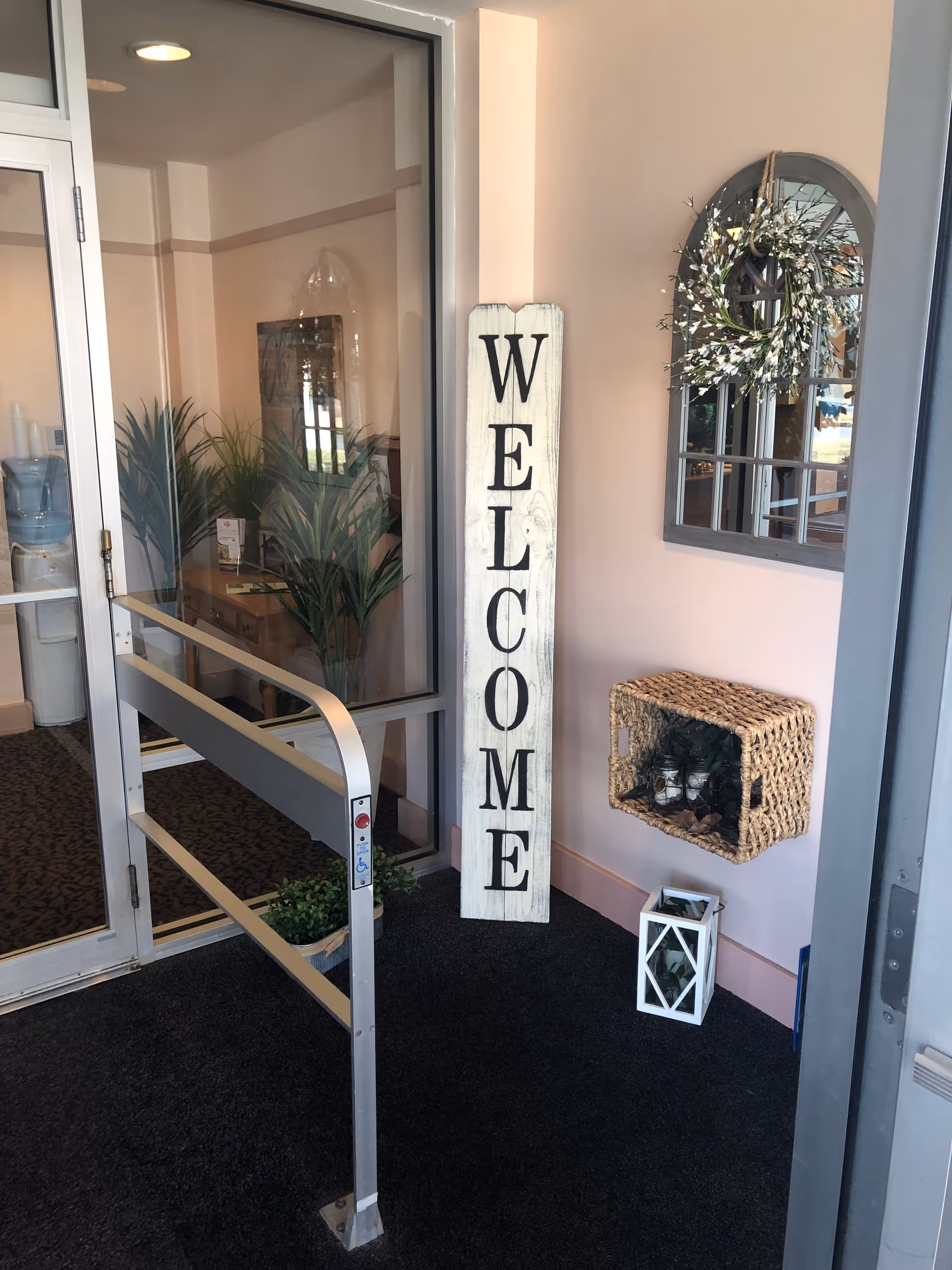 Entryway of a senior living community with a tall vertical 'WELCOME' sign, glass door and accessibility rail, potted plants, and decorative mirror and wicker shelf.