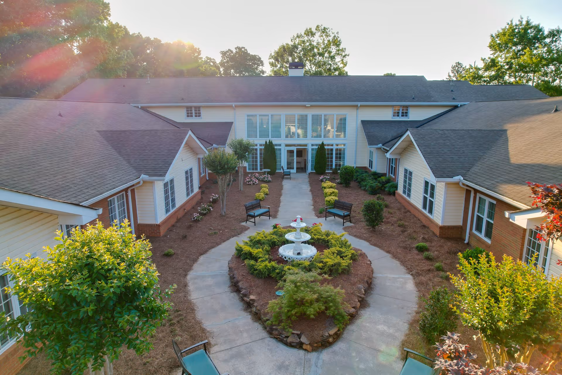 Outdoor courtyard area of a senior living facility with a central white fountain surrounded by greenery and a circular walkway. Benches are placed along the walkway, and the building with large windows and multiple entrances surrounds the courtyard. Trees and shrubs are visible, and sunlight filters through the trees.
