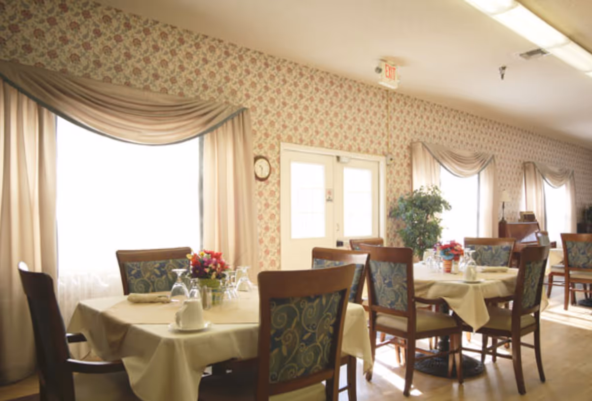 A bright dining room with several tables covered in beige tablecloths, each set with glassware, napkins, and small floral centerpieces. The room has large windows with beige curtains and floral wallpaper. Wooden chairs with patterned upholstery surround the tables, and there is a potted plant near a set of double doors.