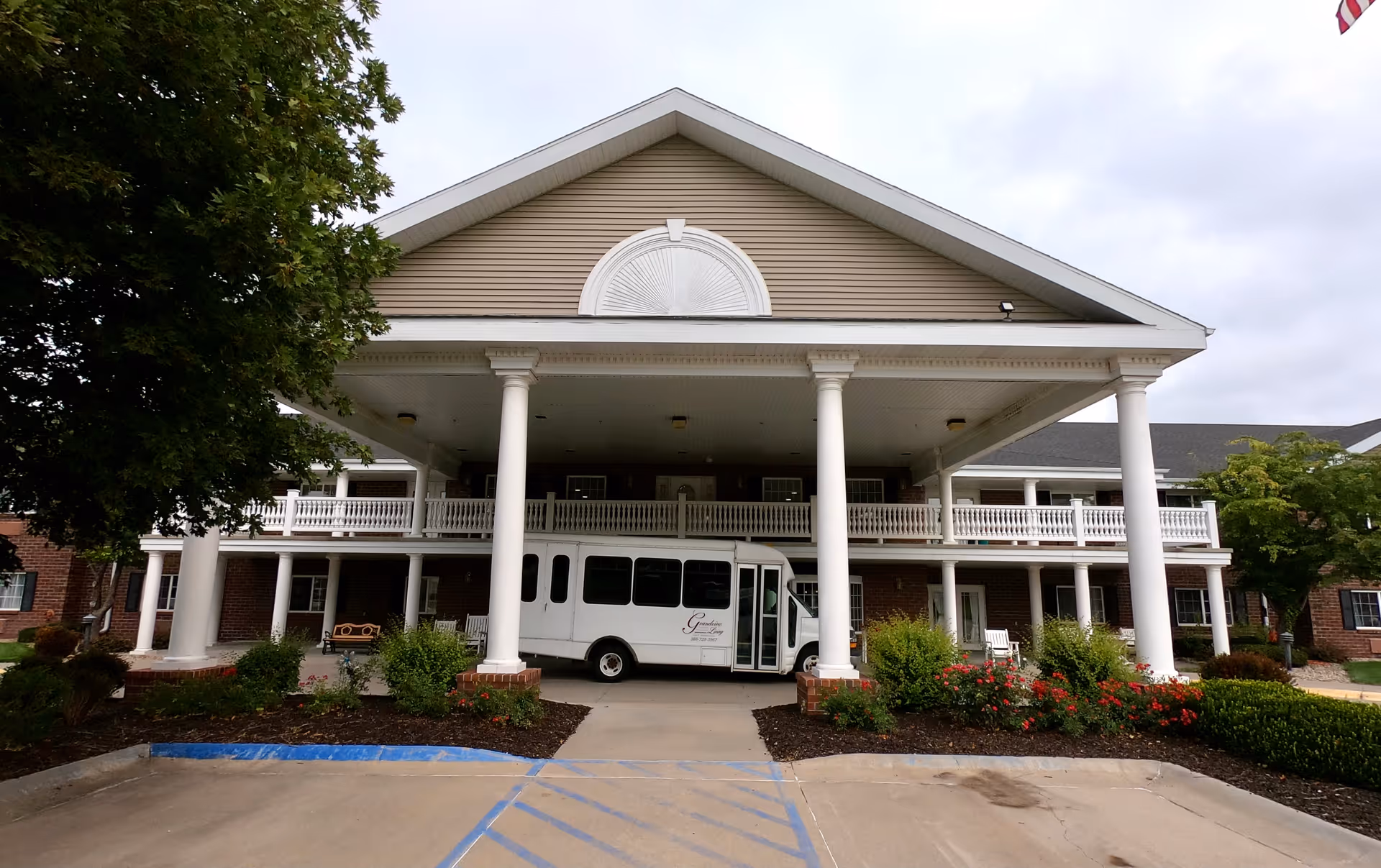 Front exterior view of a two-story assisted living facility with large white columns supporting a covered entrance. A white shuttle bus is parked under the entrance. There are bushes and flowers in front of the building, and trees on either side.