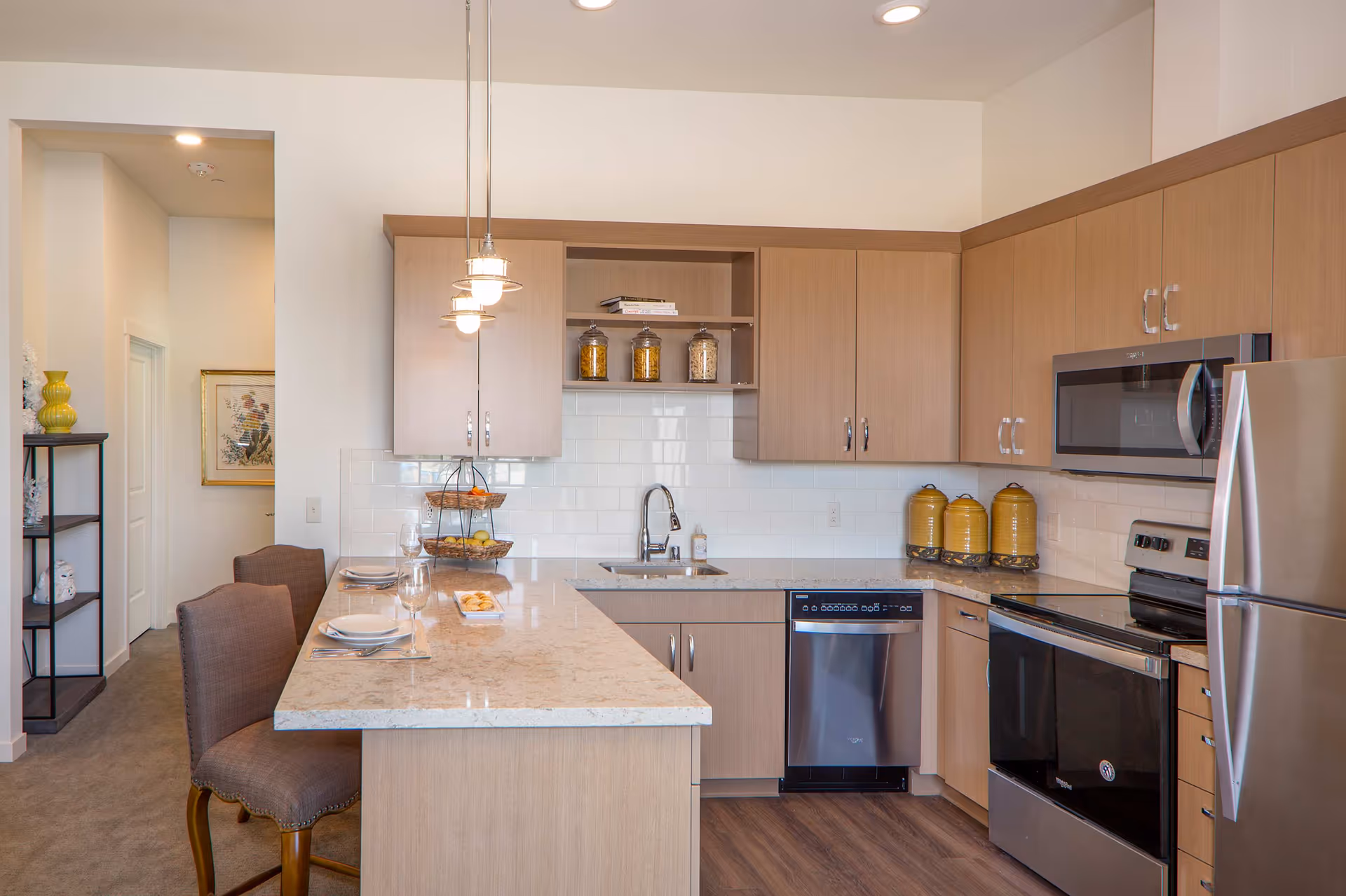 Modern kitchen with light wood cabinets, stainless steel appliances including a refrigerator, oven, microwave, and dishwasher. A marble countertop island with two upholstered chairs is set with plates, glasses, and cutlery. Three pendant lights hang above the island. There are jars with food items on the shelves and counter, and a sink with a faucet is visible.