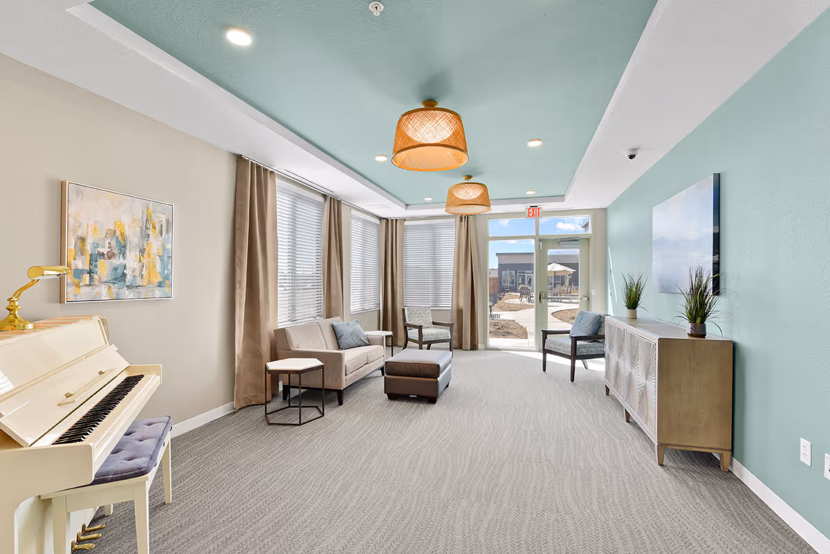 A bright and airy living room with a light blue ceiling and beige walls. The room features a white upright piano with a cushioned bench on the left, a beige sofa with a blue pillow, two armchairs, a brown ottoman, and a wooden sideboard with two potted plants. Large windows with beige curtains allow natural light to fill the space, and two woven pendant lights hang from the ceiling. A glass door at the far end leads to an outdoor patio area.