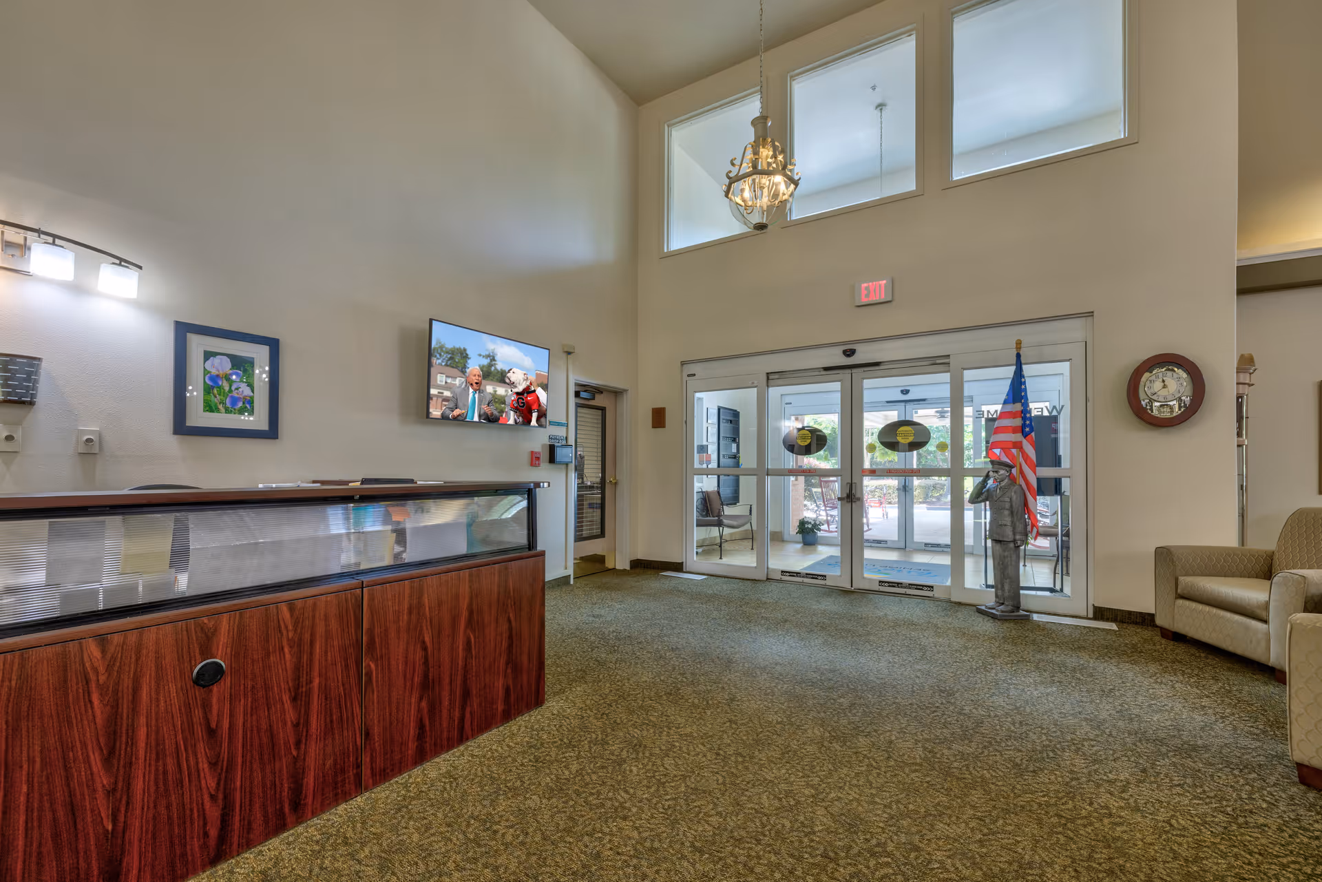 Lobby area of TerraBella Epps Bridge facility with a wooden reception desk on the left, a wall-mounted TV showing a man and a mascot, an American flag next to a statue of a saluting soldier near the glass entrance doors, beige walls, carpeted floor, and a chandelier hanging from the ceiling.
