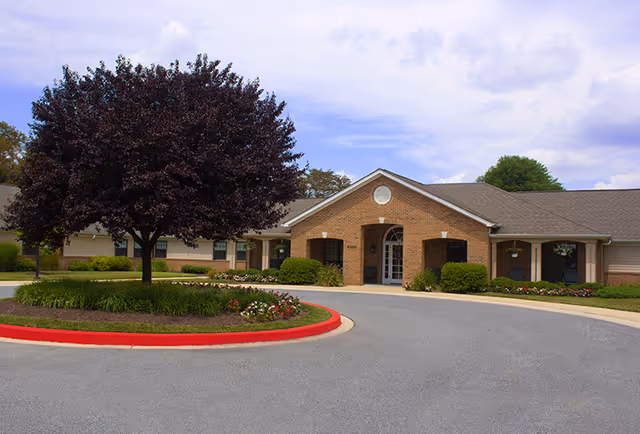Front exterior view of a single-story brick building with a gabled roof, surrounded by landscaped greenery including a tree and flower beds, under a partly cloudy sky.