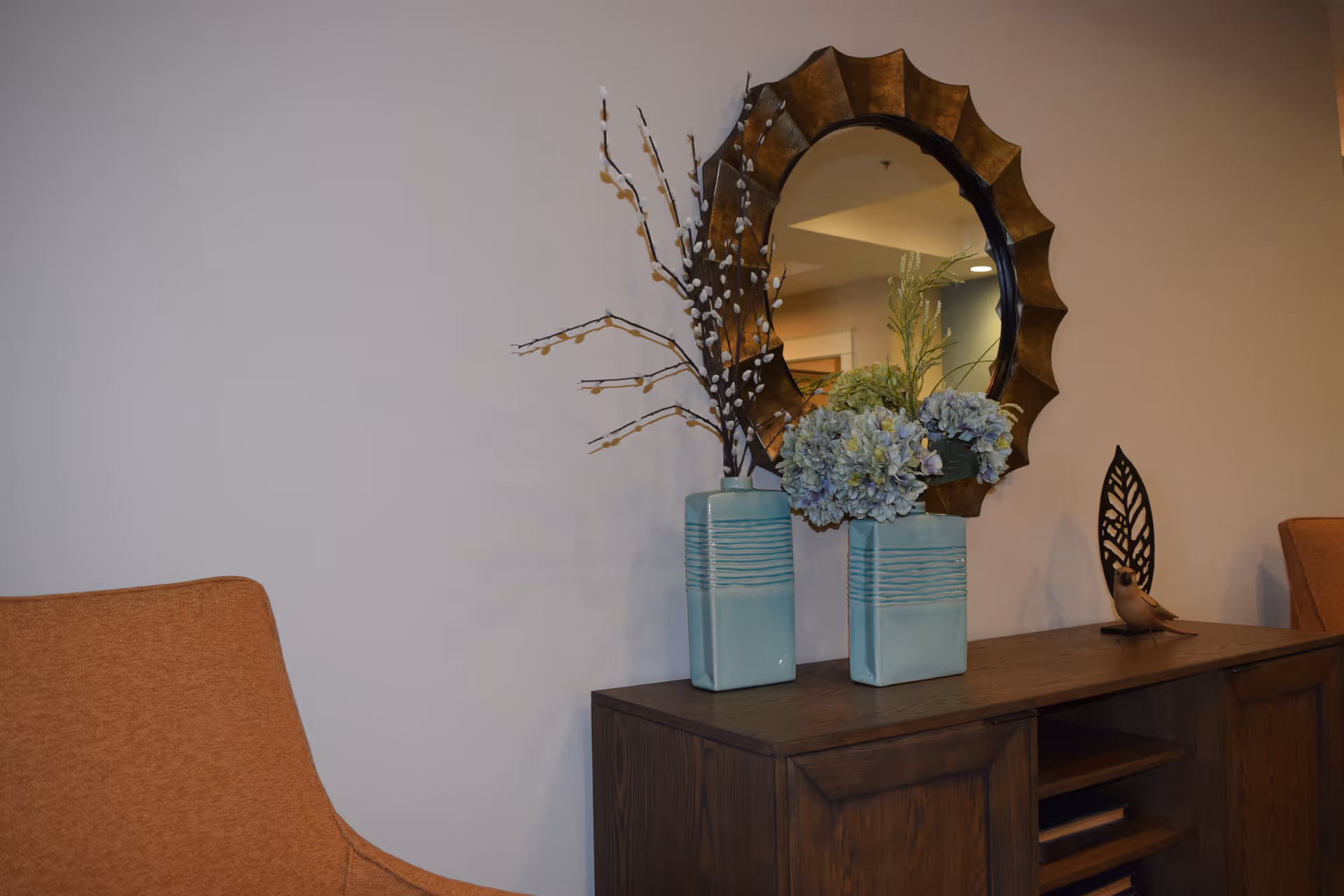 Decorative wooden console table with two blue vases of flowers, a round decorative mirror, and orange chairs against a beige wall.