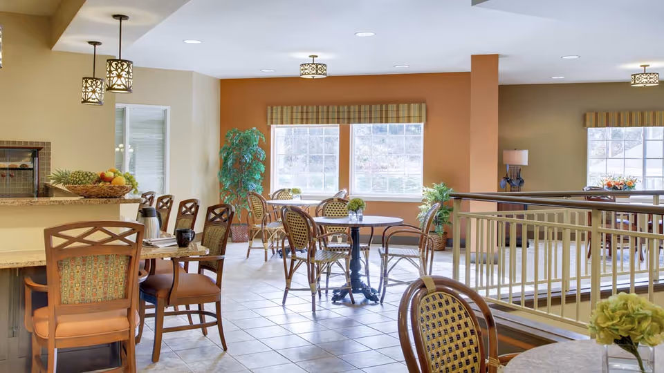 A bright and spacious dining area in The Landing of Poughkeepsie featuring multiple tables and chairs arranged for seating. The room has large windows letting in natural light, potted plants for decoration, and pendant lights hanging from the ceiling. There is a counter with a fruit basket and coffee thermos on it.