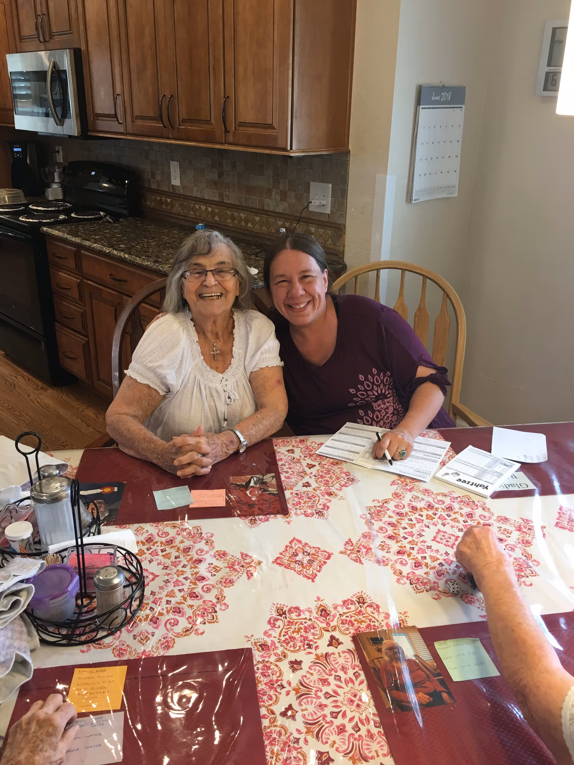 Two women sitting at a dining table in a kitchen, smiling at the camera. The table is covered with a red and white patterned tablecloth and has various papers and items on it. The kitchen has wooden cabinets, a microwave, and a stove in the background.