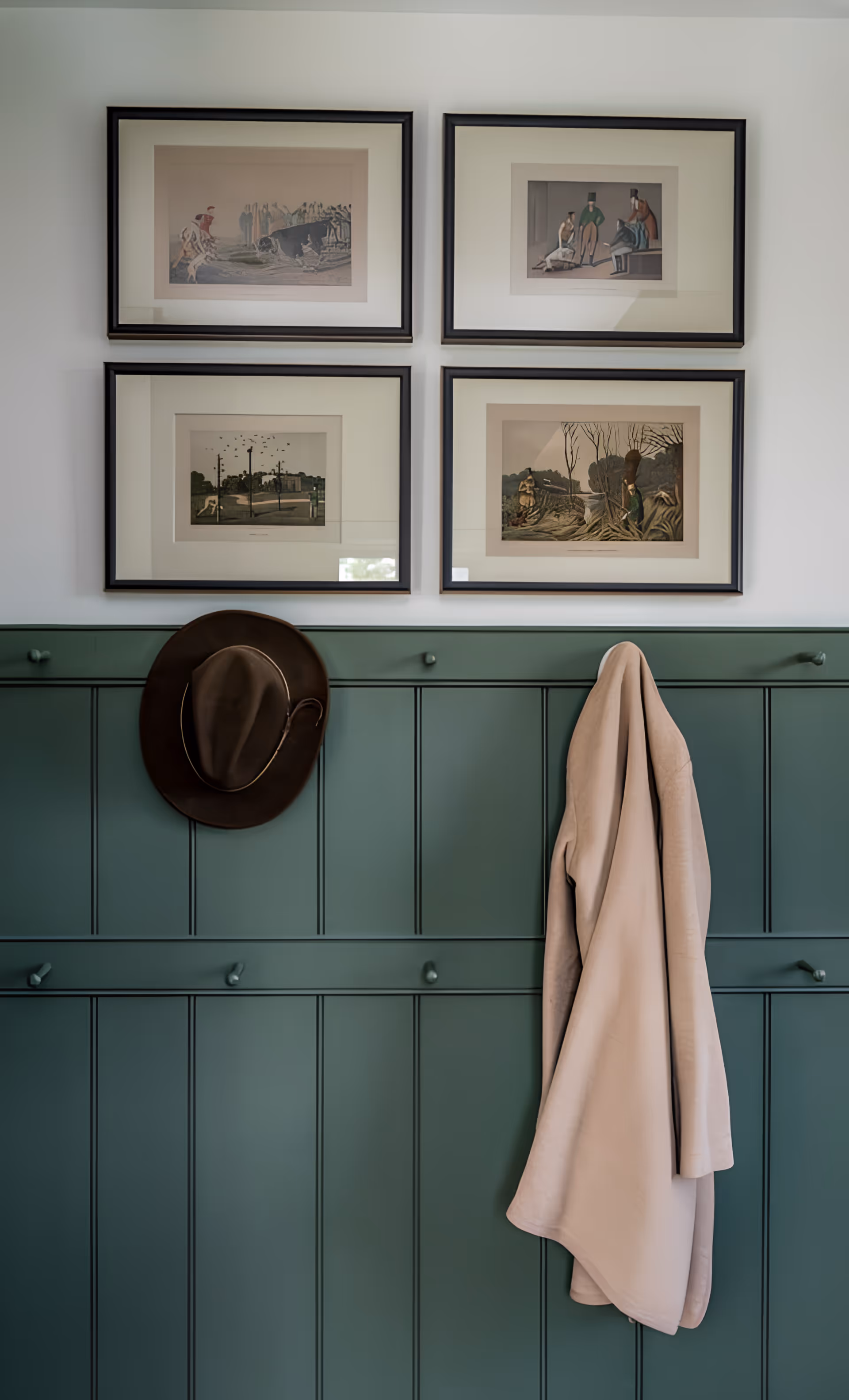 A green paneled wall with four framed vintage-style prints hanging above. A brown hat and a beige coat are hanging on hooks attached to the wall paneling.