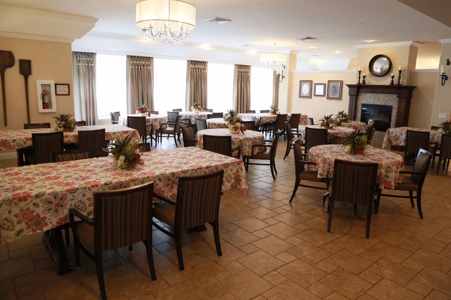 A dining room with multiple tables covered in floral tablecloths, each decorated with a small floral centerpiece. The room features large windows with curtains, a fireplace with a mirror and framed pictures above it, and chandeliers hanging from the ceiling. The floor is tiled and the chairs have striped upholstery.