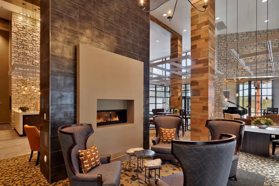A cozy and modern living room area with four upholstered armchairs arranged around a small set of nesting tables in front of a contemporary fireplace. The room features a mix of stone and wood wall accents, large windows allowing natural light, and a grand piano in the background.