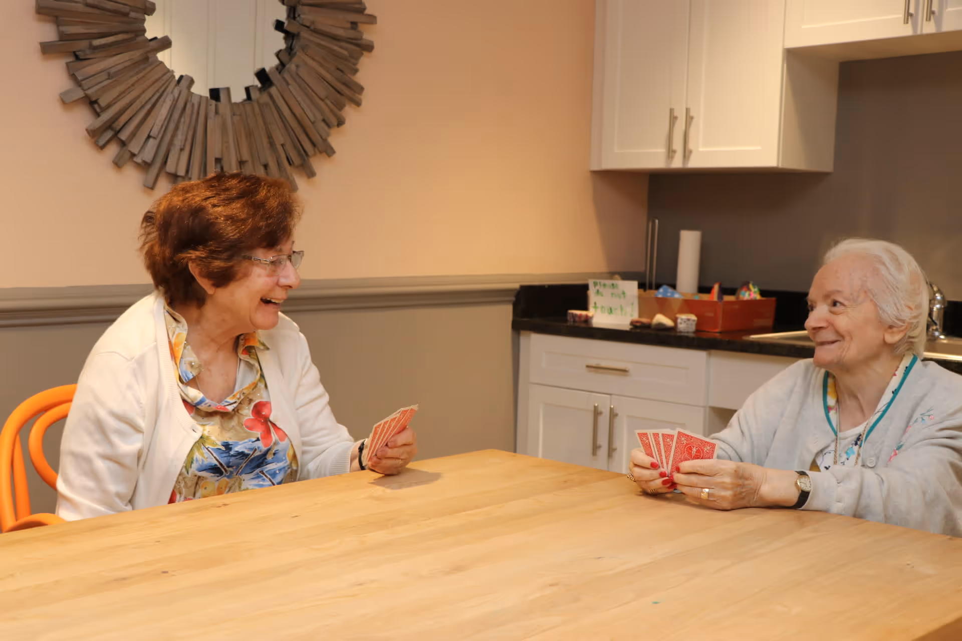 Two elderly women sitting at a wooden table playing cards and smiling at each other in a room with white cabinets and a decorative round mirror on the wall.