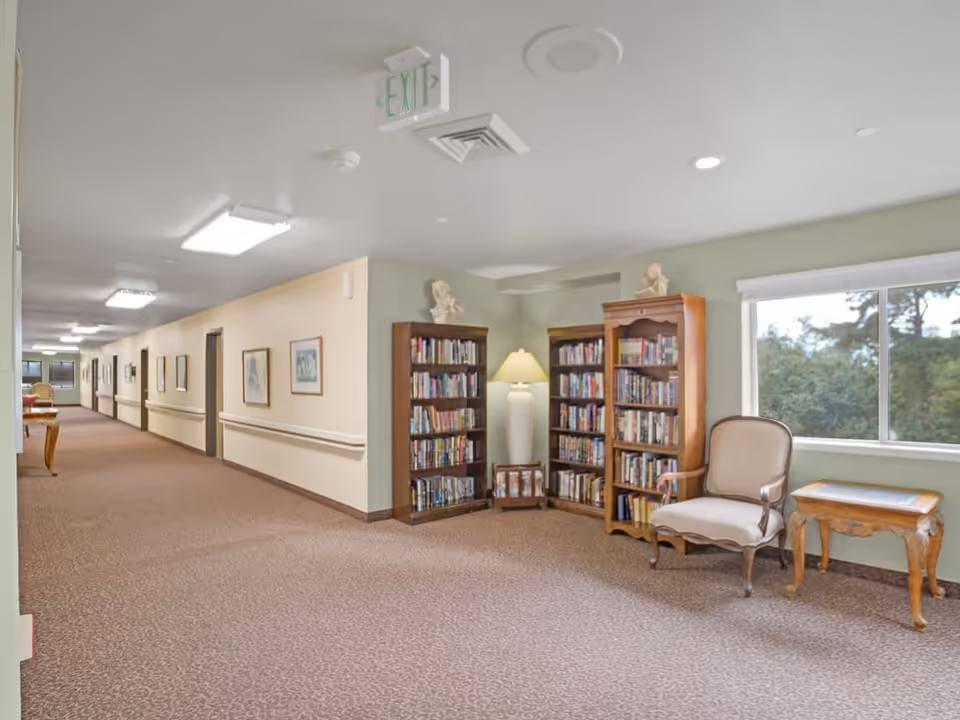 A well-lit hallway in a senior living facility with beige walls and carpeted floor. On the right side, there are two wooden bookshelves filled with books, a floor lamp between them, a cushioned armchair, and a small wooden side table next to a window showing greenery outside. The hallway extends into the distance with framed pictures on the walls and several doors.