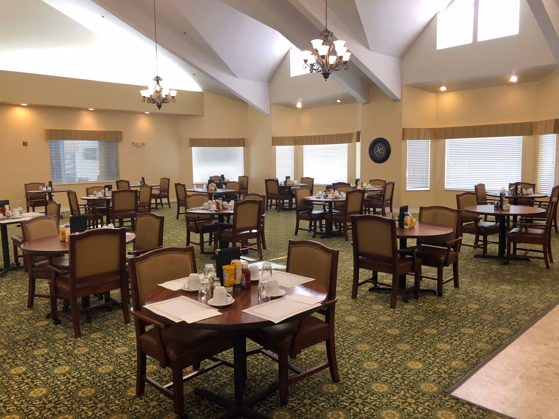 Spacious senior dining room with multiple round tables and chairs set for meals under vaulted ceilings and chandeliers.