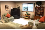 A cozy living room with light green upholstered sofas and armchairs, decorated with orange and beige throw pillows. There is a large television in the corner, a wooden bookshelf filled with books, a small wooden table with plants, and a window letting in natural light.