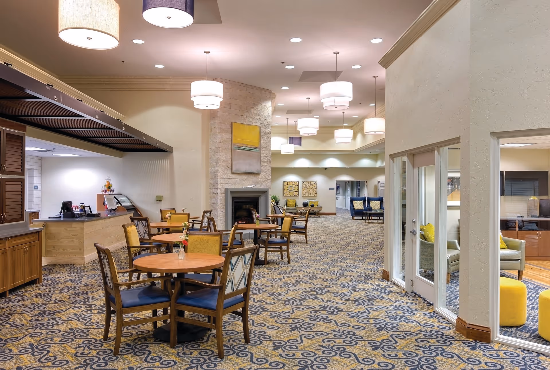 A spacious and well-lit common area in a senior living facility featuring round wooden tables with chairs, a stone fireplace with a painting above it, and multiple hanging ceiling lights. The carpet has a blue and yellow pattern, and there is a small counter area on the left side. On the right, a glass-walled room with yellow and green chairs and yellow ottomans is visible.