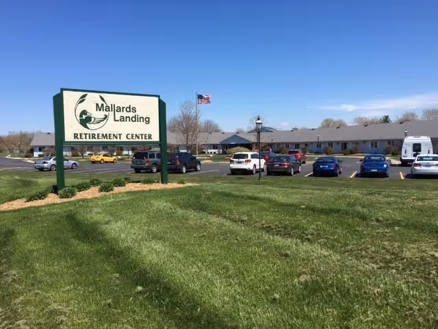 Exterior view of Mallards Landing Retirement Center with a large green and white sign in the foreground. The building is a single-story structure with a gray roof, and there are several cars parked in the parking lot. An American flag is flying on a flagpole near the building under a clear blue sky.