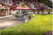 Outdoor patio area with tables and chairs on a paved surface next to a well-maintained grassy lawn. The patio is adjacent to a building with large windows and a red roof. Trees with red leaves provide partial shade.