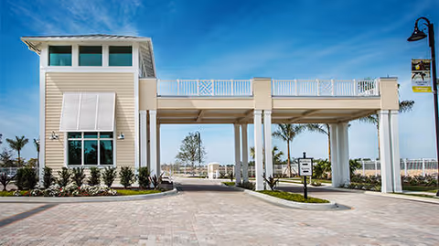 Porte-cochère entrance of a light-colored community building with columns, a paved driveway, palm trees, and a blue sky.