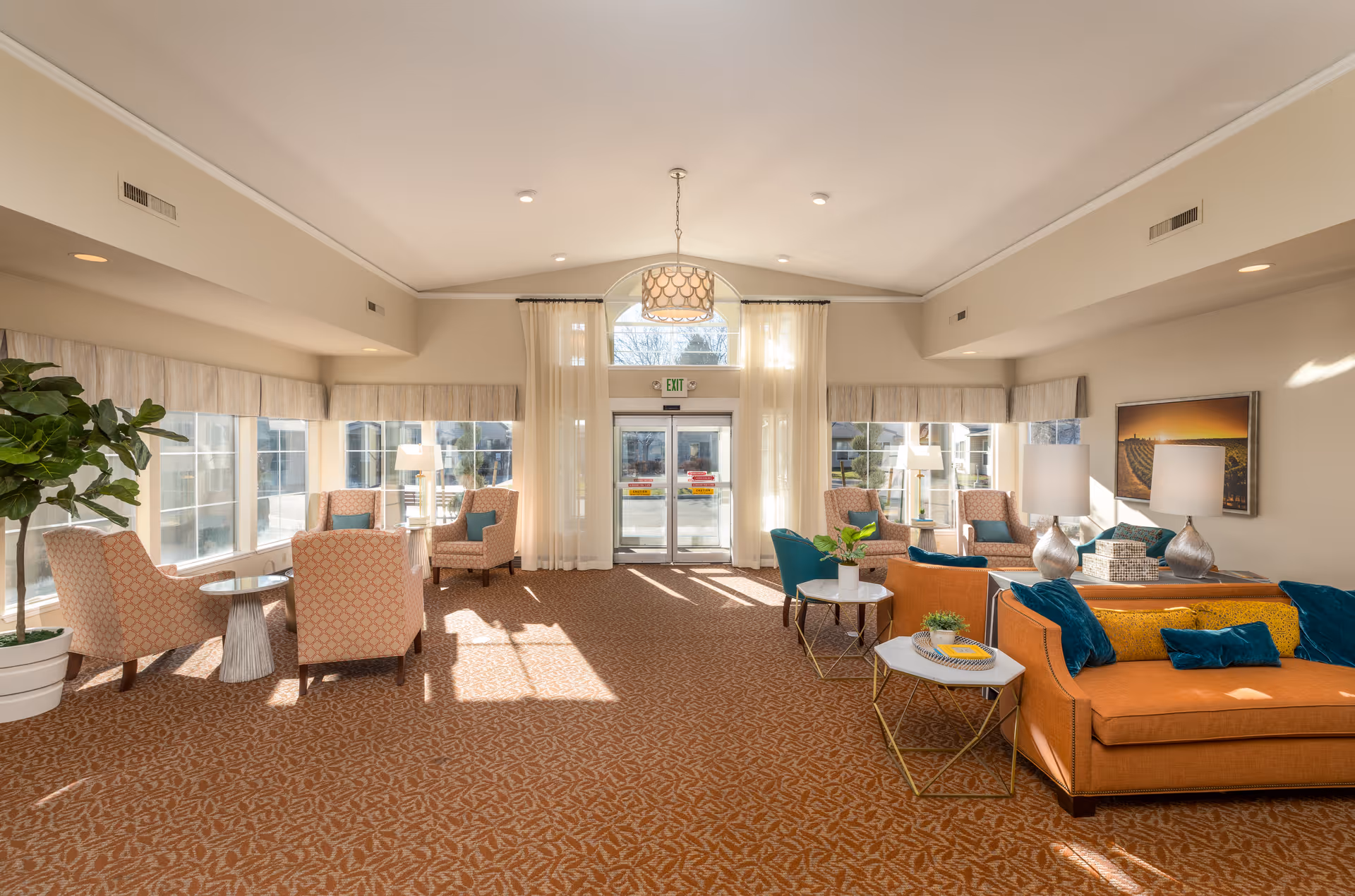 A bright and spacious living room area in a senior living facility with large windows letting in natural light. The room features multiple seating arrangements including orange sofas with blue and yellow cushions, patterned armchairs, small round tables with plants and lamps, and sheer curtains hanging over glass double doors leading outside.