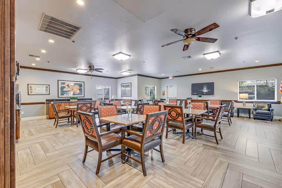 A spacious dining room with multiple square tables and chairs featuring patterned upholstery. The room has light-colored tiled flooring, ceiling fans, and recessed lighting. There are windows letting in natural light, framed artwork on the walls, and a flat-screen TV mounted on one wall. A small seating area with two black chairs and a side table is visible near the windows.