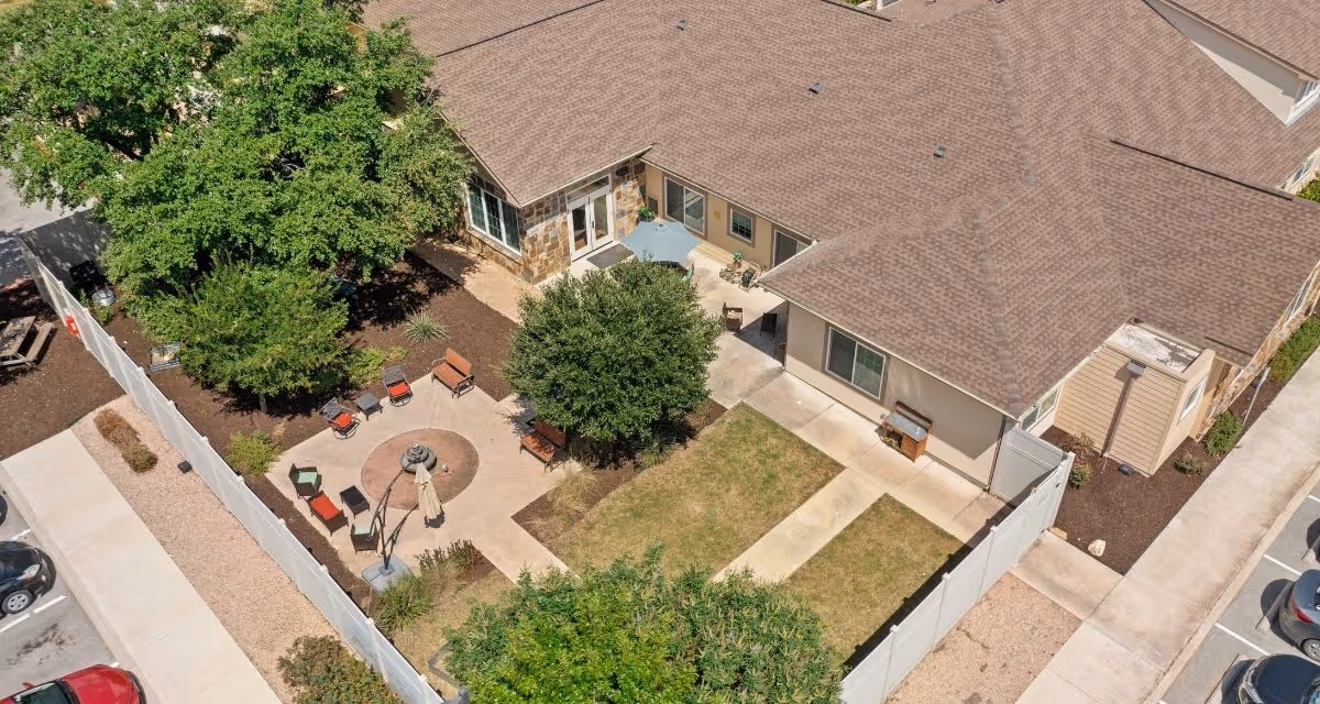 Aerial view of an outdoor courtyard area at Poet's Walk San Antonio, A Memory Care Community. The courtyard features a circular seating area with multiple chairs and a central umbrella, surrounded by trees and shrubs. The building has a brown shingled roof and beige walls, with concrete walkways and a fenced perimeter. There are parked cars visible on the left side of the image.