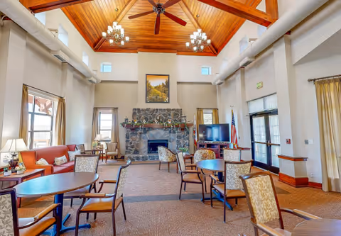 A spacious living room with high wooden vaulted ceilings, ceiling fans, and chandeliers. The room features a stone fireplace with a framed picture above it, multiple round tables with chairs, a red sofa, side tables with lamps, and a television near an American flag. Large windows and glass doors allow natural light to fill the space.