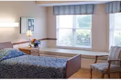 A bright senior bedroom with a single bed, bedside table and lamp, window seat beneath large windows, and an upholstered chair.