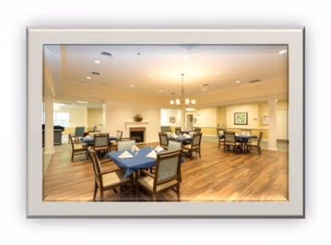 A spacious dining room with wooden floors and several tables covered with blue tablecloths. Each table is surrounded by chairs, and some tables have folded napkins placed on them. The room is well-lit with ceiling lights and a chandelier. There is a fireplace on one wall and framed artwork hanging on the walls.