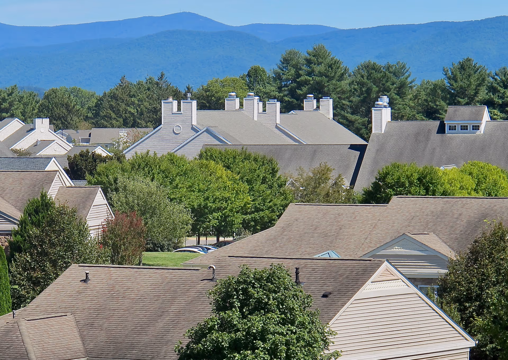 View of multiple residential buildings with beige roofs and light-colored siding surrounded by green trees, with a backdrop of blue mountains under a clear sky.