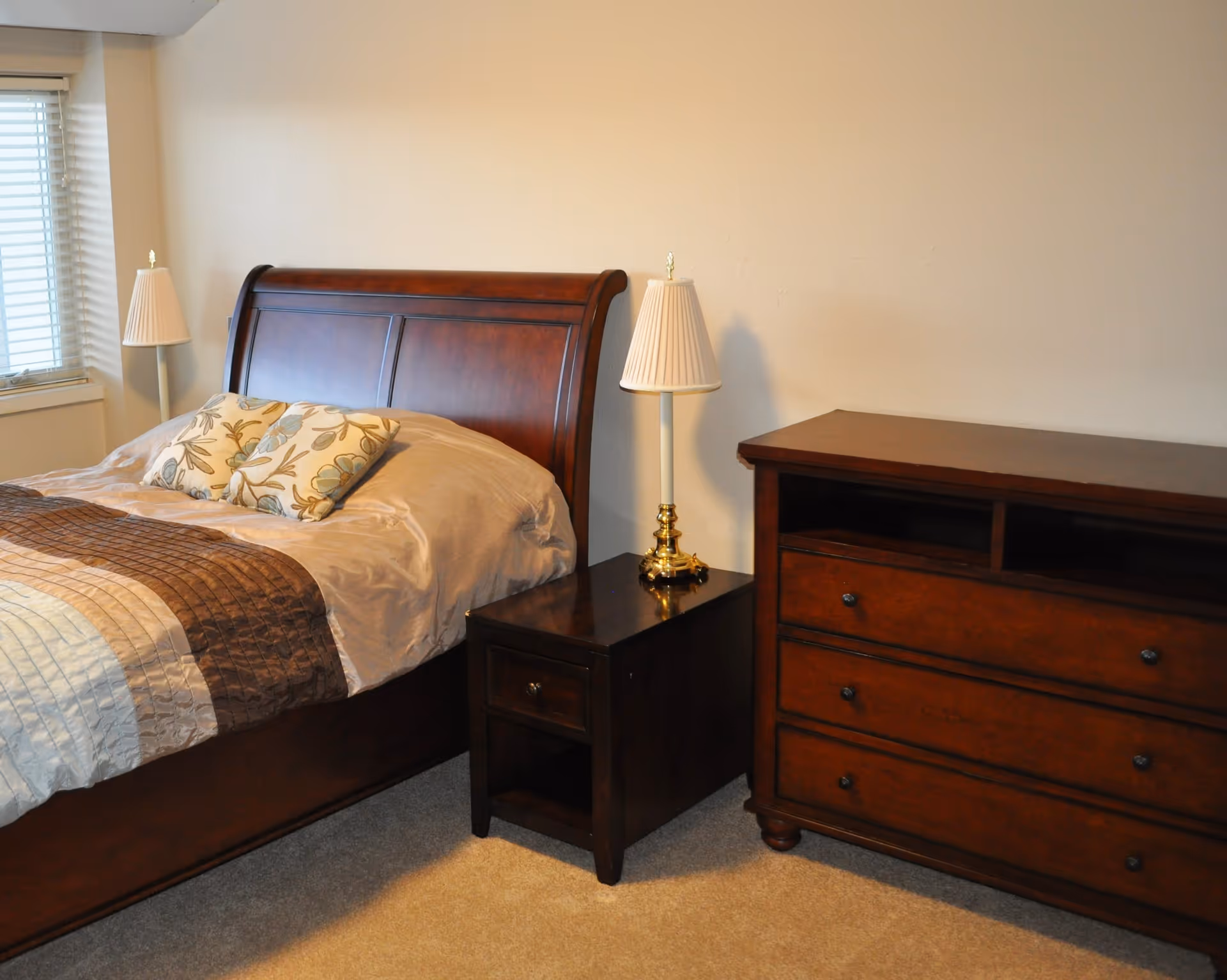 A bedroom with a wooden bed frame, beige and brown bedding, two floral pillows, a wooden nightstand with a brass lamp, and a wooden dresser against a beige wall. A window with blinds is visible on the left side.