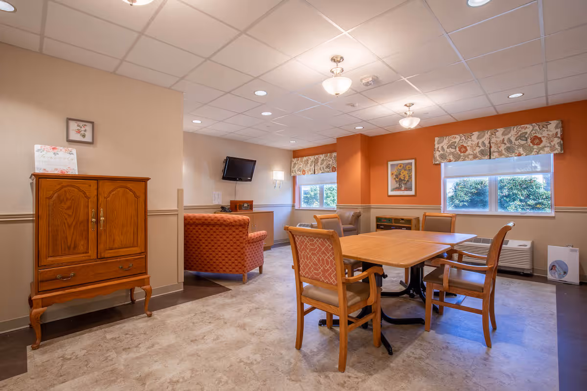 A cozy common area in a nursing and rehabilitation center featuring a wooden table with four chairs, an orange armchair, a wooden cabinet, two windows with floral valances, a wall-mounted TV, and warm orange and beige walls.