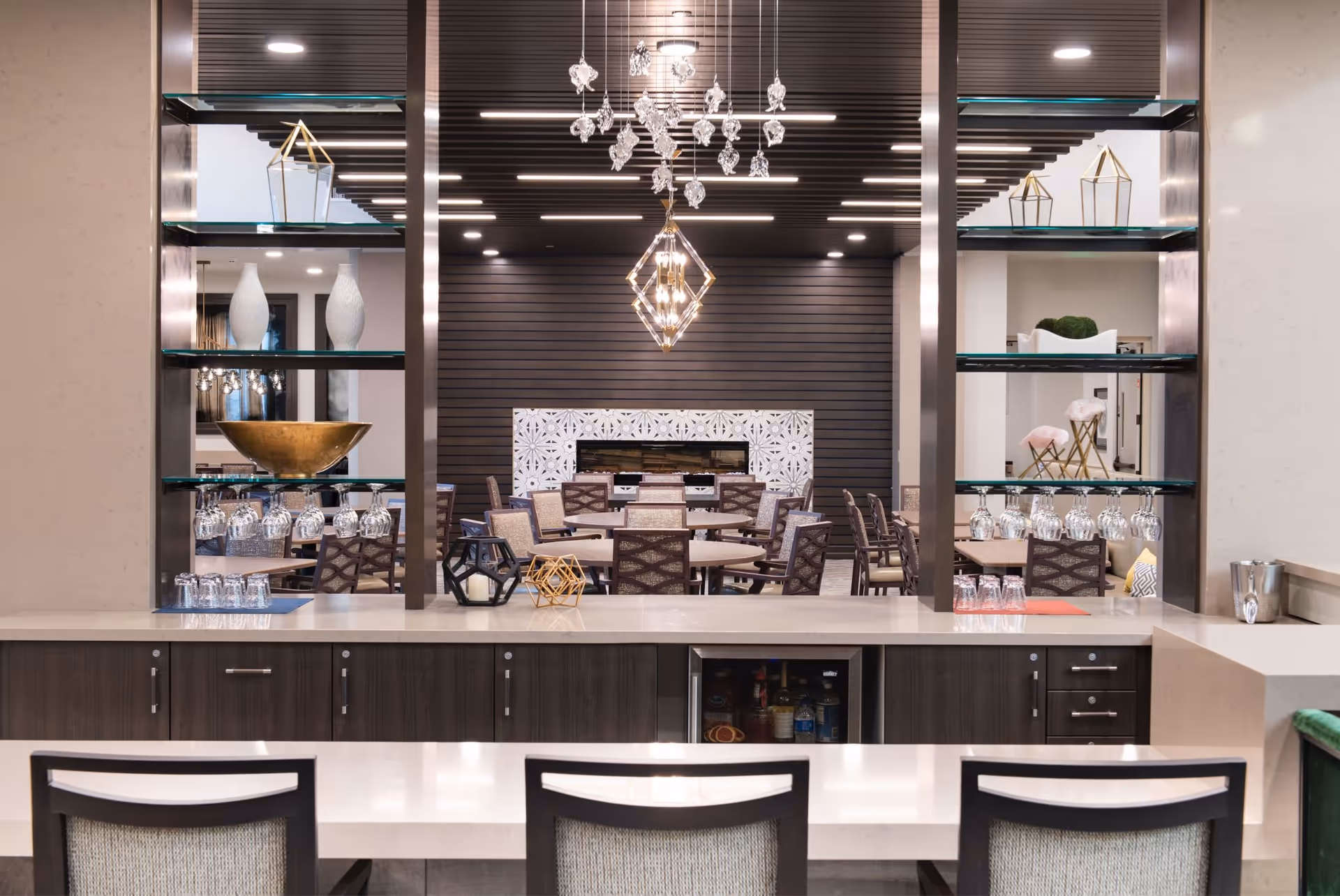 Modern communal dining room with a bar counter and chairs in the foreground, glass shelving, chandeliers, and tables and chairs in the background.