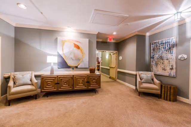 A well-lit hallway area in a senior living facility with beige carpet and gray walls. The space features two beige armchairs with cushions on either side of a wooden sideboard with geometric patterns. A modern table lamp and decorative items are on the sideboard. Two abstract paintings hang on the walls, and an open door leads to another hallway with handrails. Ceiling lights illuminate the area.