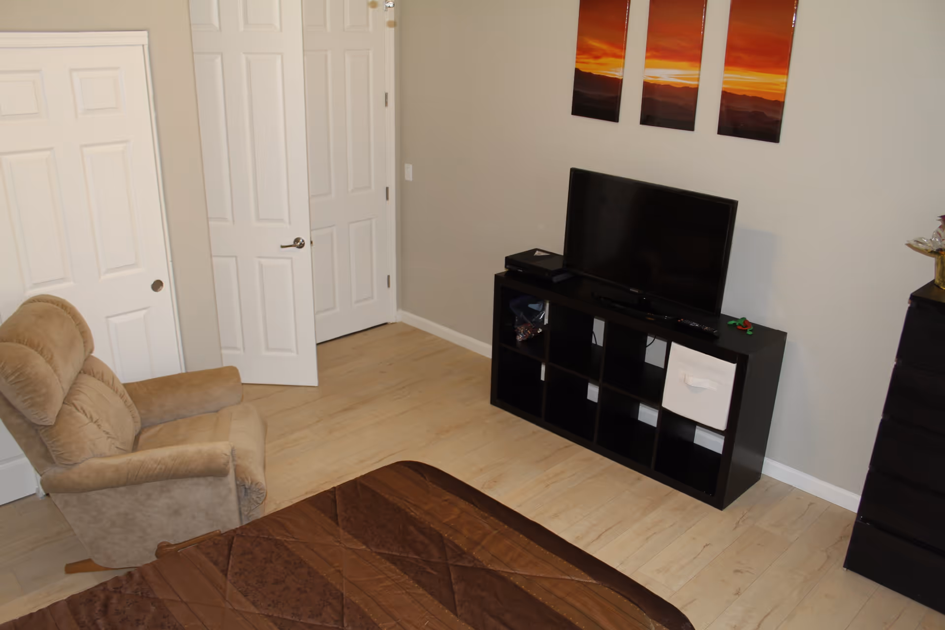 A modest bedroom with a bed in the foreground, a recliner to the left, and a TV on a black shelving unit against a light-colored wall.