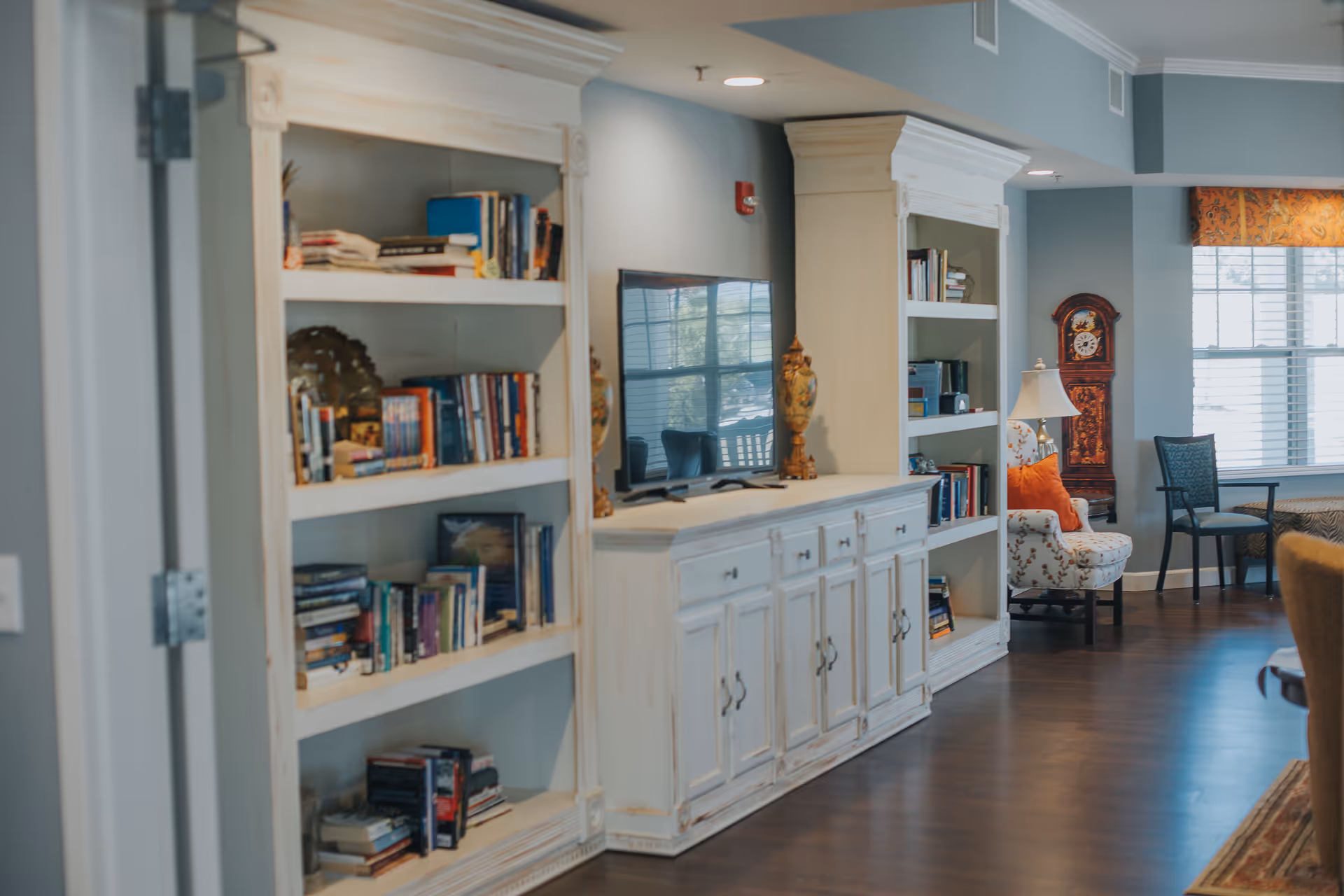 Interior view of a senior living facility common area featuring white built-in bookshelves filled with books, a flat-screen TV on a white cabinet, a decorative clock, a floral armchair with an orange pillow, and a window with blinds and a patterned valance.
