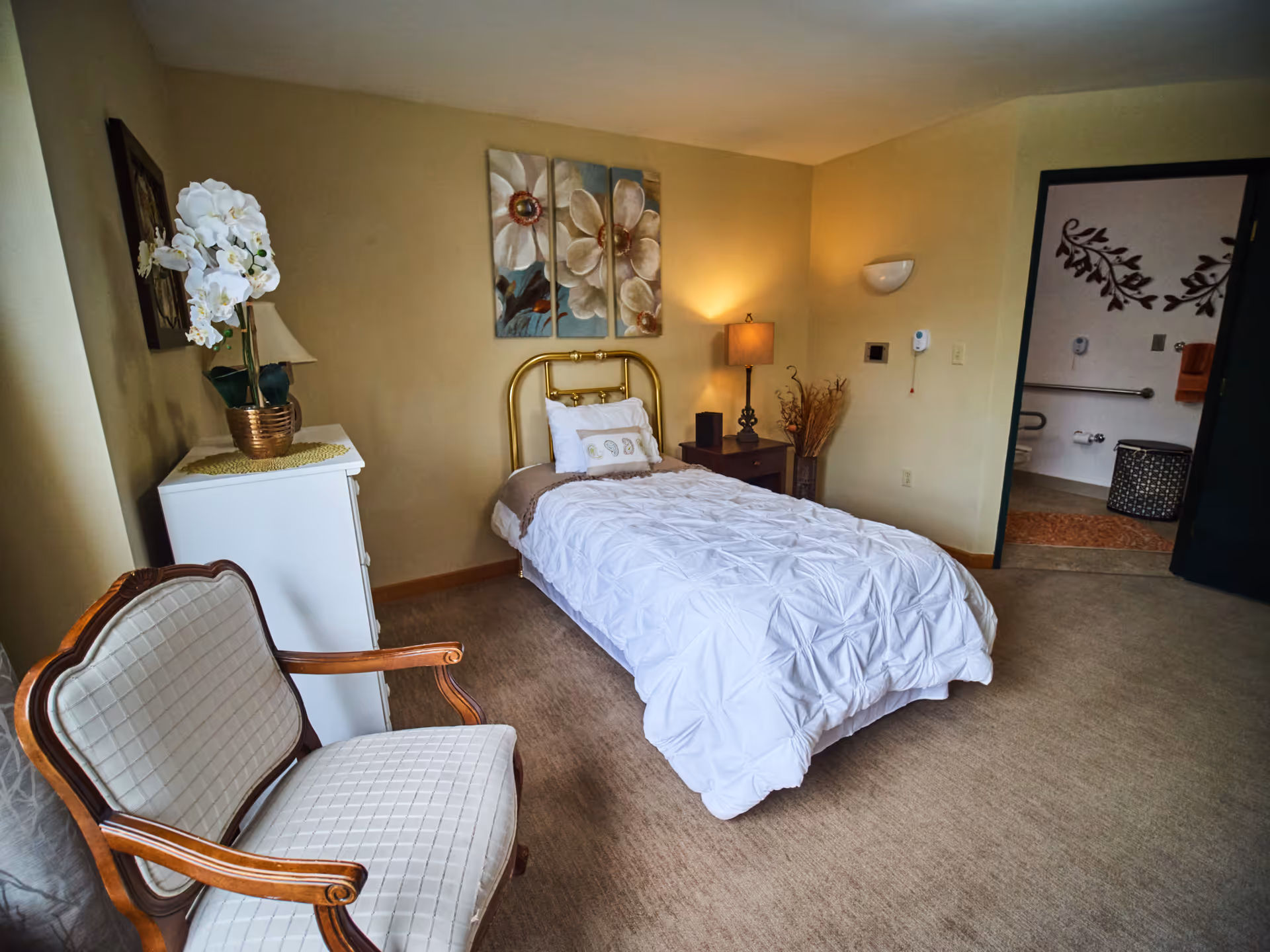 A cozy senior living bedroom with a single bed featuring a white comforter and decorative pillows. There is a wooden nightstand with a lamp and decorative dried plants beside the bed. A white dresser with a potted white orchid sits against one wall, and a cushioned wooden armchair is positioned near the dresser. The room has beige walls with floral artwork above the bed. An open door reveals a bathroom with grab bars and a laundry basket.
